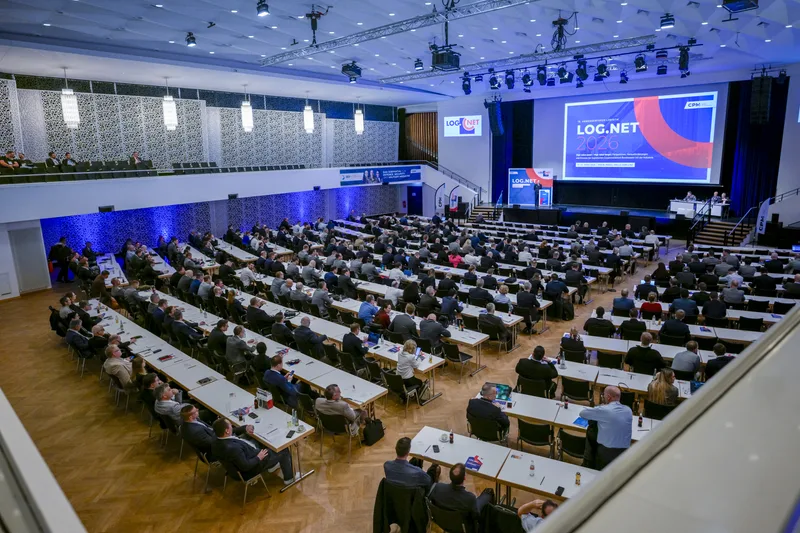 Blick in eine gut besuchte Veranstaltungshalle während einer Fachtagung. Die Teilnehmer sitzen an langen Tischreihen und richten ihren Blick auf die Bühne, auf der ein Redner vor einer großen Leinwand mit einer Veranstaltungsankündigung spricht.