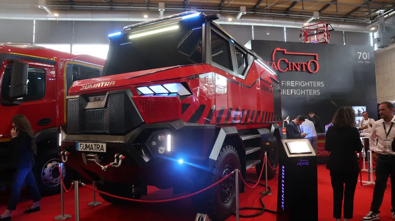 Ein modernes, rot-schwarzes Feuerwehrfahrzeug des Typs „Sumatra“ ist auf einer Fachmesse ausgestellt. Im Hintergrund des Messestands sind mehrere Besucher sowie Werbewände des Herstellers zu sehen.