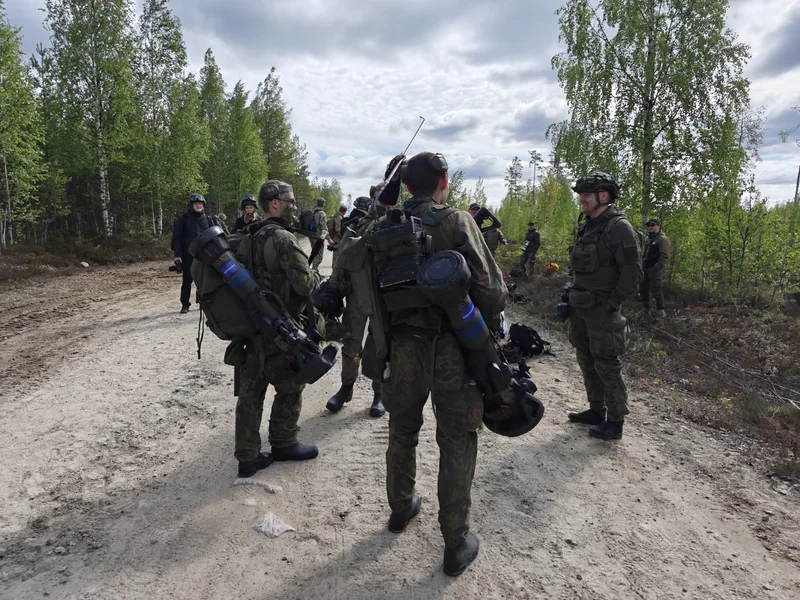 Mehrere Soldaten in militärischer Tarnkleidung stehen auf einem unbefestigten Waldweg und unterhalten sich. Zwei von ihnen tragen schultergestützte Panzerabwehrwaffen mit blauen Markierungen auf dem Rücken, während im Hintergrund weitere Personen in einem bewaldeten Gebiet erkennbar sind.