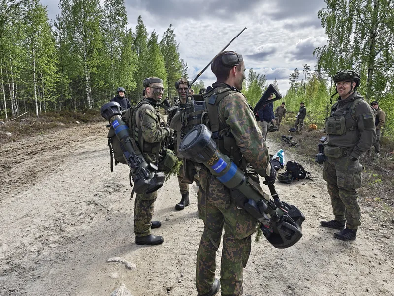 Mehrere Soldaten in Tarnkleidung stehen auf einem unbefestigten Waldweg und führen ein Gespräch. Zwei von ihnen tragen schultergestützte Panzerabwehrwaffen auf dem Rücken, während ein Soldat auf der rechten Seite einen Kennzeichnungsaufnäher für Medienvertreter an seiner Ausrüstung trägt.