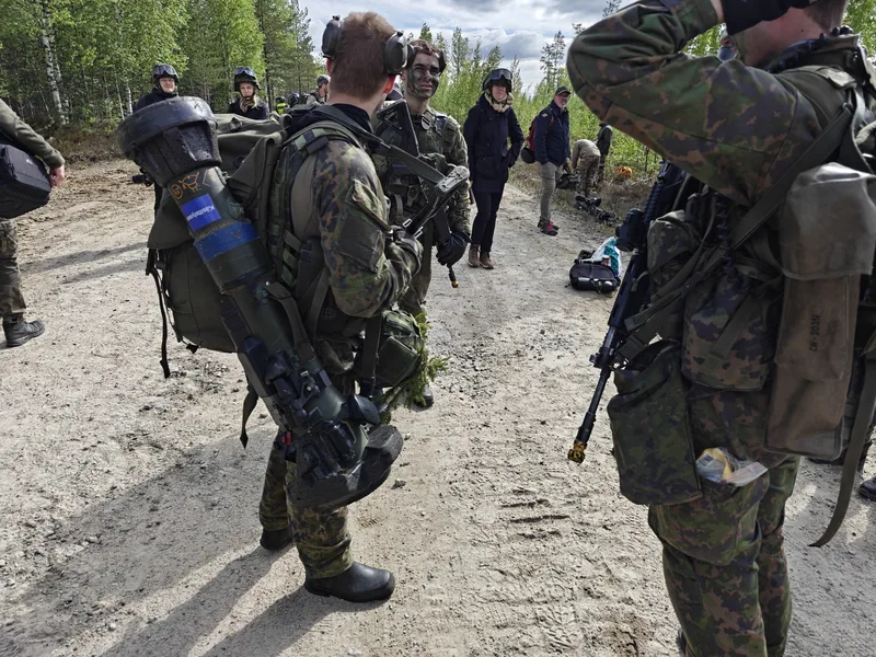 Mehrere Soldaten in Tarnkleidung stehen auf einem unbefestigten Weg in einem Waldgebiet. Ein Soldat im Vordergrund trägt eine Panzerabwehrwaffe mit einer blauen Kennzeichnung für Übungsgeräte auf dem Rücken, während andere Soldaten mit Sturmgewehren ausgerüstet sind. Im Hintergrund sind weitere Personen, teils mit Helmen, zu sehen.