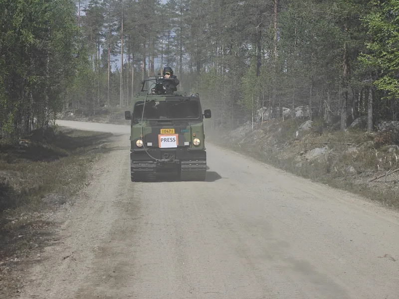 Abgebildet ist ein militärisches Kettenfahrzeug, das auf einem staubigen Waldweg fährt. An der Front des Fahrzeugs ist ein Schild zur Kennzeichnung von Medienvertretern angebracht, während eine Person mit Helm aus der Dachluke blickt.