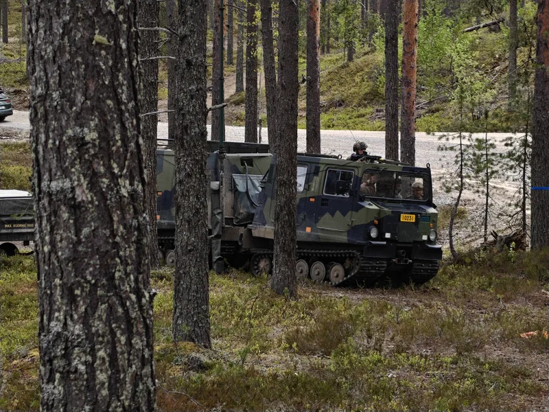 Abgebildet ist ein getarntes, zweigeteiltes Kettenfahrzeug mit Soldaten in einem lichten Nadelwald. Ein Soldat sichert aus der geöffneten Dachluke des Fahrzeugs, während im Hintergrund ein unbefestigter Weg verläuft.