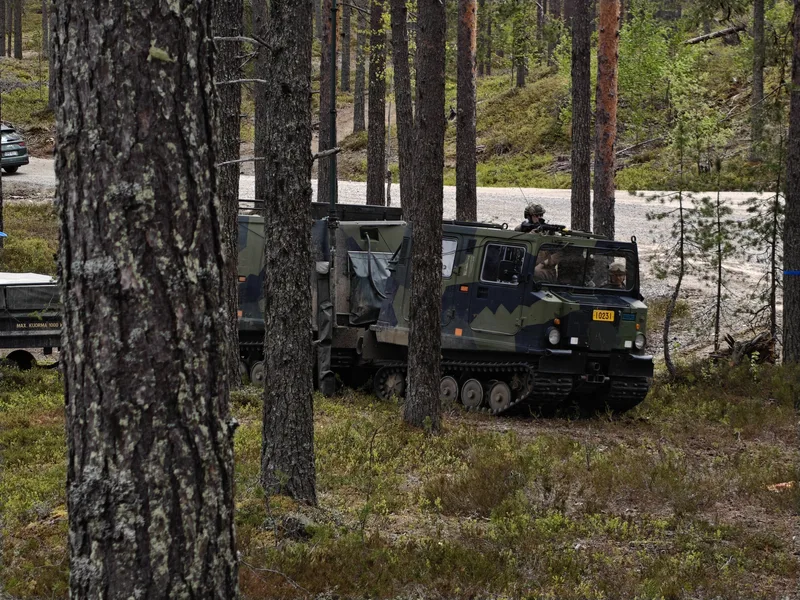 Abgebildet ist ein getarntes, geländegängiges Kettenfahrzeug mit Soldaten, das sich in einem lichten Nadelwald befindet. Ein Soldat sichert die Umgebung aus der Dachluke des Fahrzeugs, an das ein Anhänger mit einer Aufschrift zur maximalen Traglast angekoppelt ist.