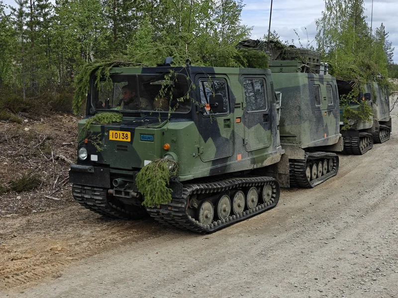 Zwei mit Tarnbemalung und Zweigen getarnte militärische Kettenfahrzeuge stehen hintereinander auf einem unbefestigten Weg am Waldrand. Im Führerhaus des vorderen Fahrzeugs, das mit einem gelben Kennzeichen versehen ist, ist eine Person am Steuer erkennbar.
