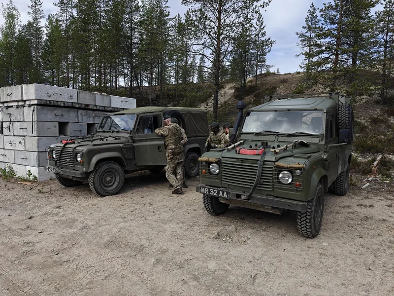 Zwei olivgrüne militärische Geländewagen stehen neben gestapelten Betonblöcken auf einem sandigen Gelände vor einem Waldstück. Mehrere Soldaten in Tarnkleidung und grünen Baretten halten sich bei den Fahrzeugen auf.