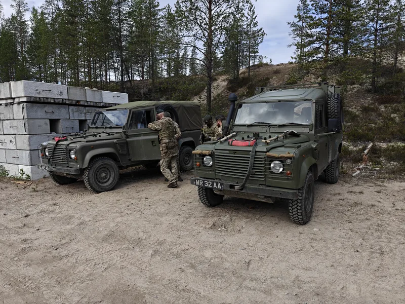 Zwei olivgrüne militärische Geländewagen stehen auf einem sandigen Gelände vor einem Waldstück. Drei Soldaten in Tarnkleidung und mit Baretten halten sich bei den Fahrzeugen auf, während sich auf der linken Seite gestapelte Betonblöcke befinden.