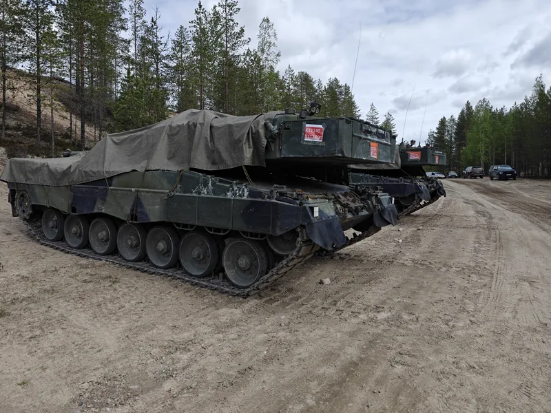 Abgebildet sind zwei Kampfpanzer mit Planen über den Türmen, die auf einem unbefestigten Waldweg abgestellt sind. Auf der Rückseite der Panzertürme sind rote Aufkleber mit Kennnummern angebracht.