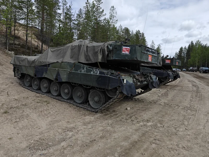 Zwei mit Planen abgedeckte Kampfpanzer stehen auf einem unbefestigten Sandweg am Rande eines Waldgebiets. An den Türmen der Fahrzeuge sind rote Kennzeichnungsaufkleber angebracht.