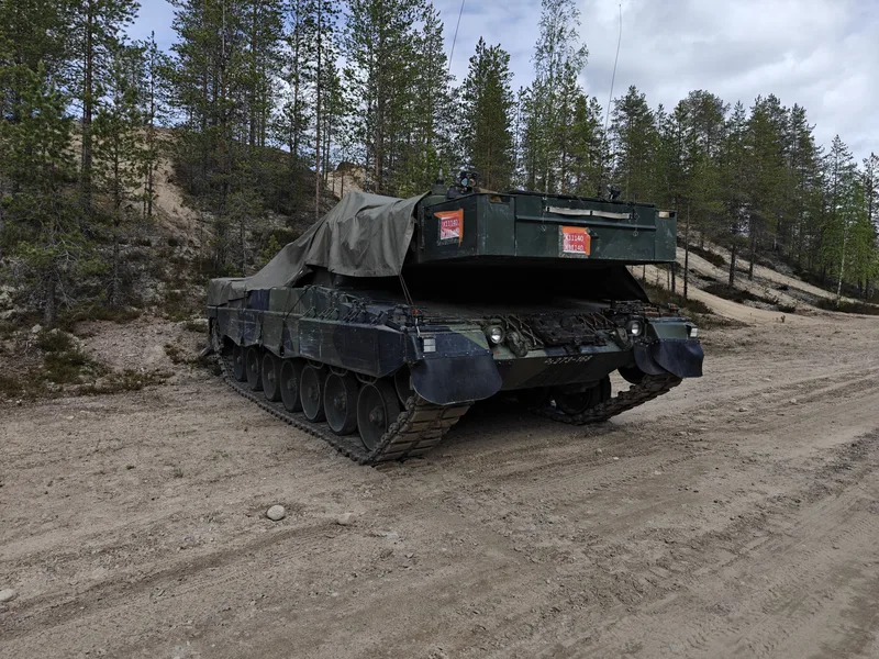 Abgebildet ist ein gepanzertes Kettenfahrzeug auf einem sandigen Weg vor einer bewaldeten Kulisse. Der obere Bereich des Fahrzeugs ist teilweise mit einer olivgrünen Plane abgedeckt, zudem befinden sich am Heck rote Kennzeichnungen.