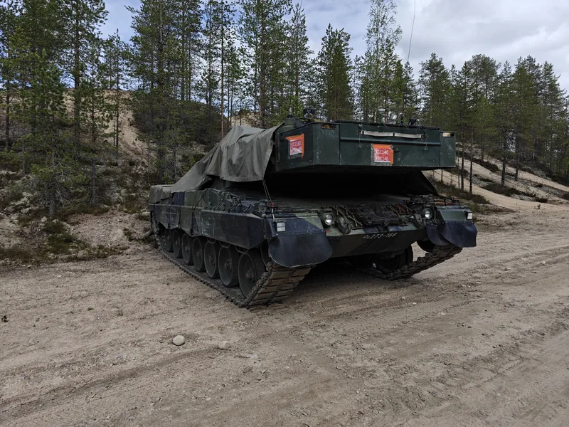Abgebildet ist ein militärisches Kettenfahrzeug, das auf einem unbefestigten Waldweg abgestellt ist. Der obere Bereich des Fahrzeugs ist teilweise mit einer olivgrünen Plane abgedeckt, zudem sind am Heck verschiedene Kennzeichnungen angebracht.