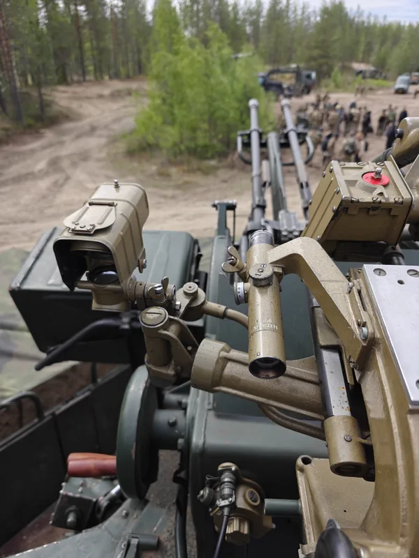 Abgebildet ist die optische Visiereinrichtung einer doppelläufigen Flugabwehrkanone im Vordergrund. Im Hintergrund sind Personen in militärischer Ausrüstung sowie Fahrzeuge auf einem unbefestigten Waldweg erkennbar.