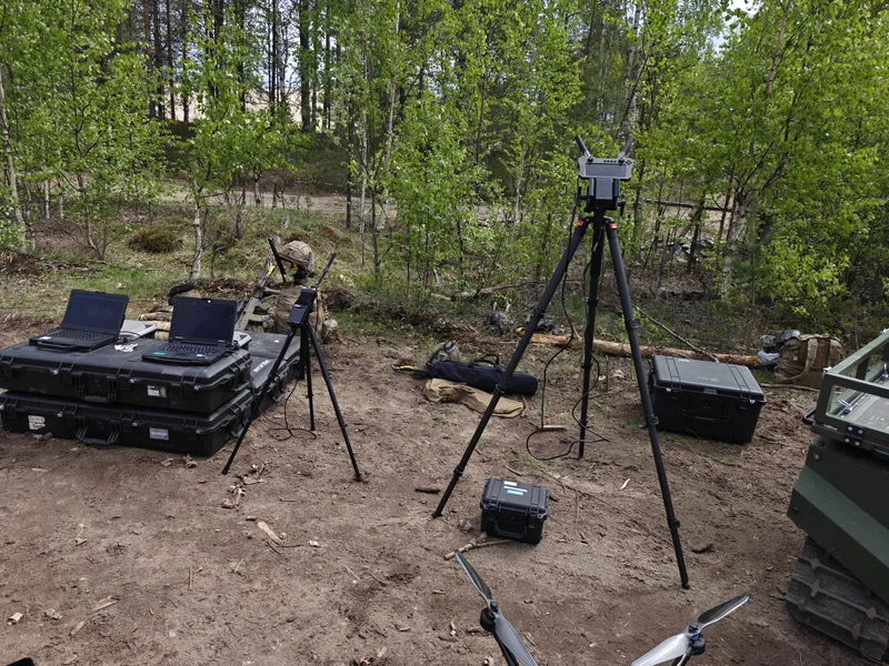 Abgebildet ist im Wald aufgebaute militärische Ausrüstung mit tragbaren Rechnern auf Transportkisten sowie Sende- und Steuerungseinheiten auf Stativen. Im Hintergrund befindet sich ein Soldat mit Gewehr, während am rechten Bildrand ein unbemanntes Kettenfahrzeug und im Vordergrund Teile einer Drohne zu sehen sind.