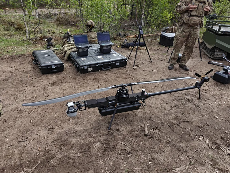 Abgebildet ist ein unbemanntes Hubschraubersystem auf unbefestigtem Boden in einem Waldgebiet. Im Hintergrund befinden sich zwei robuste Bediengeräte auf Transportkisten, eine Antenne auf einem Stativ, ein Soldat in Tarnkleidung sowie ein unbemanntes Kettenfahrzeug.