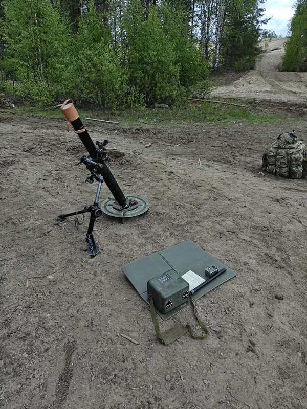 Abgebildet ist ein auf sandigem Boden im Bereich eines Waldrandes in Stellung gebrachter Mörser mit Zweibein und Bodenplatte. Neben der Waffe liegen auf einer grünen Faltmatte diverses Zubehör, darunter ein Transportbehälter und eine Tabelle, während im Hintergrund ein Rucksack in Tarnmuster abgestellt ist.