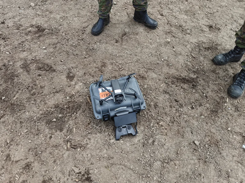 Abgebildet ist eine kleine Aufklärungsdrohne samt Fernsteuerung, die auf einem grauen Transportkoffer auf erdigem Boden bereitgestellt ist. Flankiert wird die Ausrüstung von den Beinen zweier Personen in militärischer Tarnkleidung und Stiefeln.