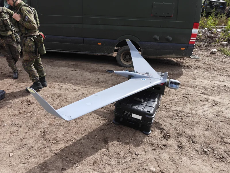Abgebildet ist eine graue Aufklärungsdrohne, die auf einem beschrifteten Transportbehälter neben einem olivgrünen Transporter abgestellt ist. Im linken Bildbereich stehen zwei Soldaten in Flecktarn-Uniformen auf unbefestigtem Boden.