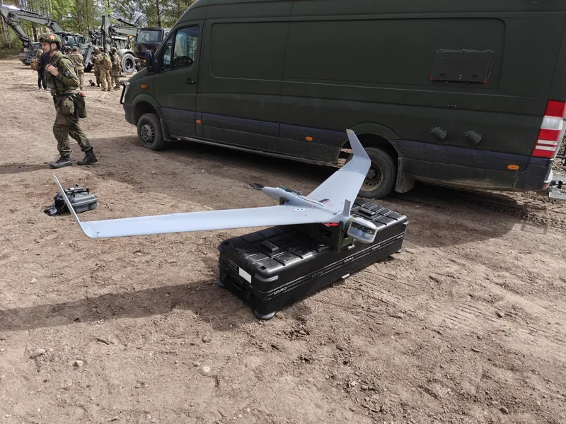 Abgebildet ist ein graues unbemanntes Luftfahrzeug, das auf einer schwarzen Transportkiste vor einem olivgrünen Kastenwagen abgestellt ist. Im Hintergrund befinden sich Soldaten in Tarnkleidung sowie militärische Baufahrzeuge auf einem unbefestigten Gelände.