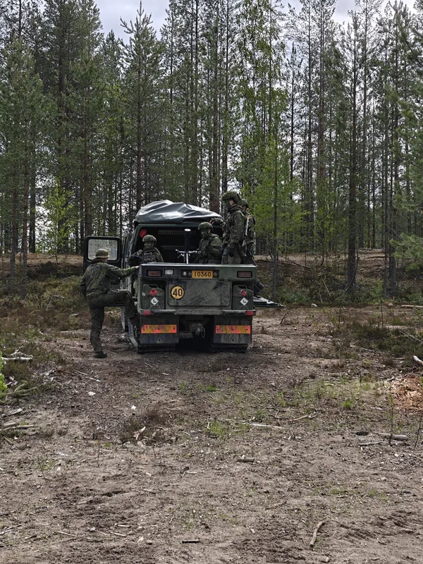 Mehrere Soldaten in Tarnkleidung befinden sich an und auf einem militärischen Kettenfahrzeug in einem Waldgebiet. Auf der Ladefläche des mit einem Kennzeichen versehenen Fahrzeugs ist ein Mörser montiert, während ein Soldat seitlich am Heck aufsteigt.
