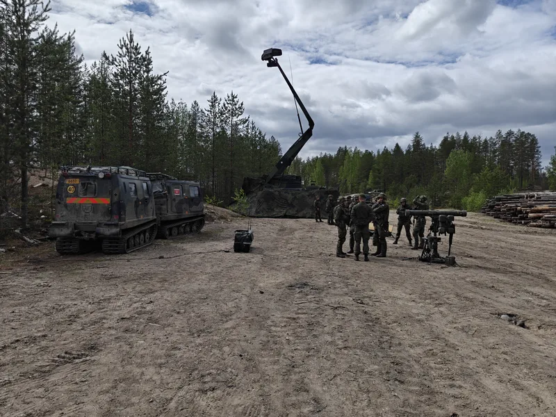 Abgebildet sind mehrere Soldaten in militärischer Ausrüstung auf einer Lichtung im Wald neben einem auf einem Stativ montierten Flugabwehrsystem. Im Hintergrund stehen ein zweigliedriges Kettenfahrzeug sowie ein mobiles Radarsystem mit weit ausgefahrenem Mast.