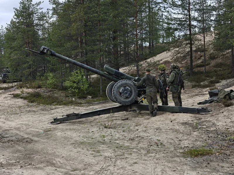 Drei Soldaten in militärischer Tarnkleidung stehen an einem in Stellung gebrachten, gezogenen Artilleriegeschütz in einem sandigen Waldgebiet. Im linken Hintergrund ist ein gepanzertes Kettenfahrzeug zwischen den Bäumen erkennbar.