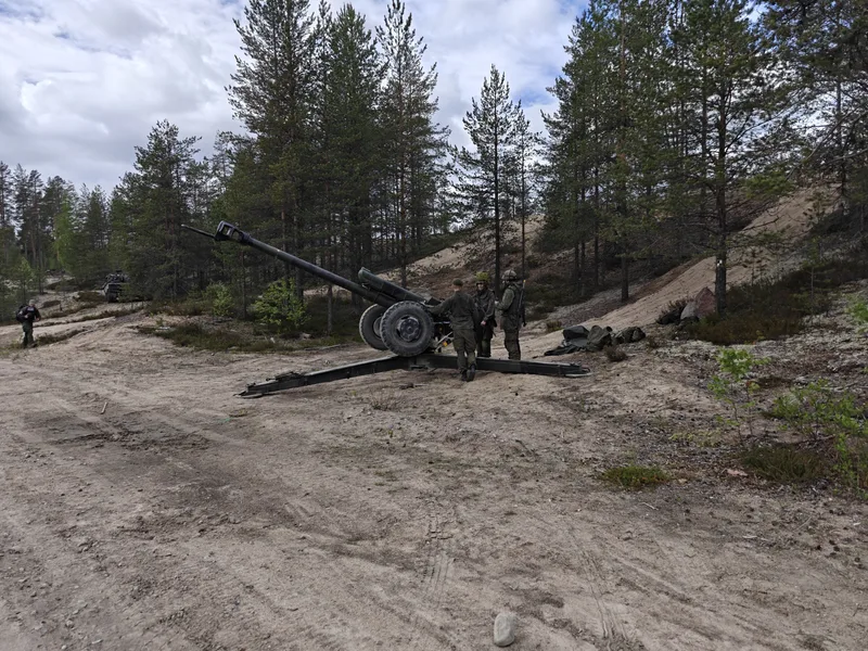 Drei Soldaten stehen an einem gezogenen Artilleriegeschütz auf einem sandigen Übungsgelände in einem Waldgebiet. Im Hintergrund sind ein weiteres Militärfahrzeug sowie eine Person zu Fuß erkennbar.