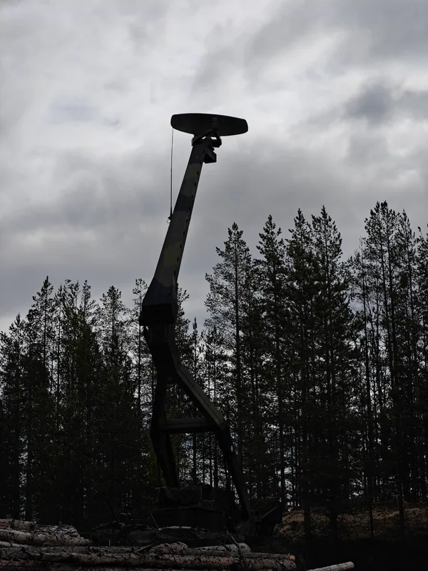 Abgebildet ist eine mobile militärische Radaranlage mit aufgerichtetem Mast vor einem Nadelwald unter bewölktem Himmel. Das getarnte System ist im Gelände positioniert, im Vordergrund sind abgelegte Baumstämme zu sehen.