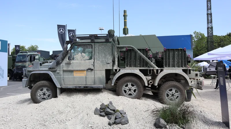 Ein dreiachsiges, geländegängiges Militärfahrzeug mit Verschmutzungsspuren steht auf einer künstlich angelegten Sandfläche im Außenbereich einer Ausstellung. Das Fahrzeug weist ein geschlossenes Führerhaus mit einem gelben Hinweisaufkleber an der Tür sowie Transportvorrichtungen auf der hinteren Ladefläche auf.