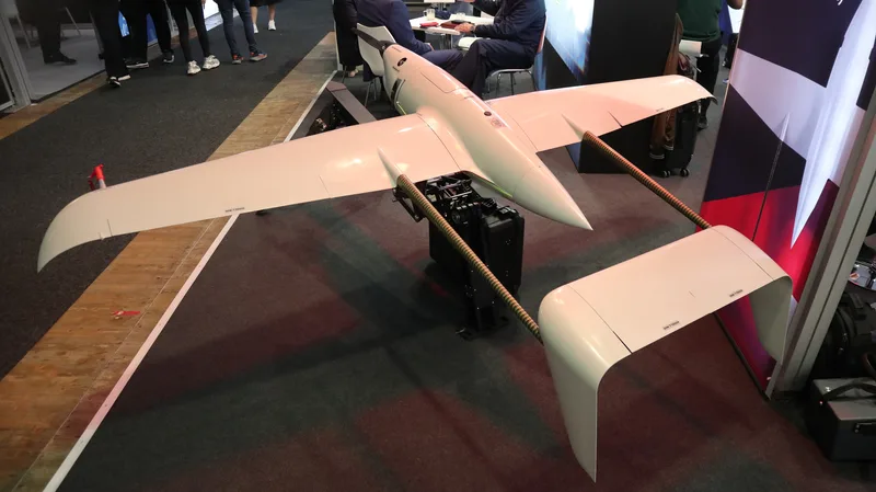 Abgebildet ist ein weißes unbemanntes Luftfahrzeug auf einem Ausstellungsstand bei einer Fachmesse. Das Fluggerät ist auf einer Halterung montiert und verfügt über einen Propellerantrieb sowie ein doppeltes Leitwerk, während im Hintergrund Messebesucher zu sehen sind.