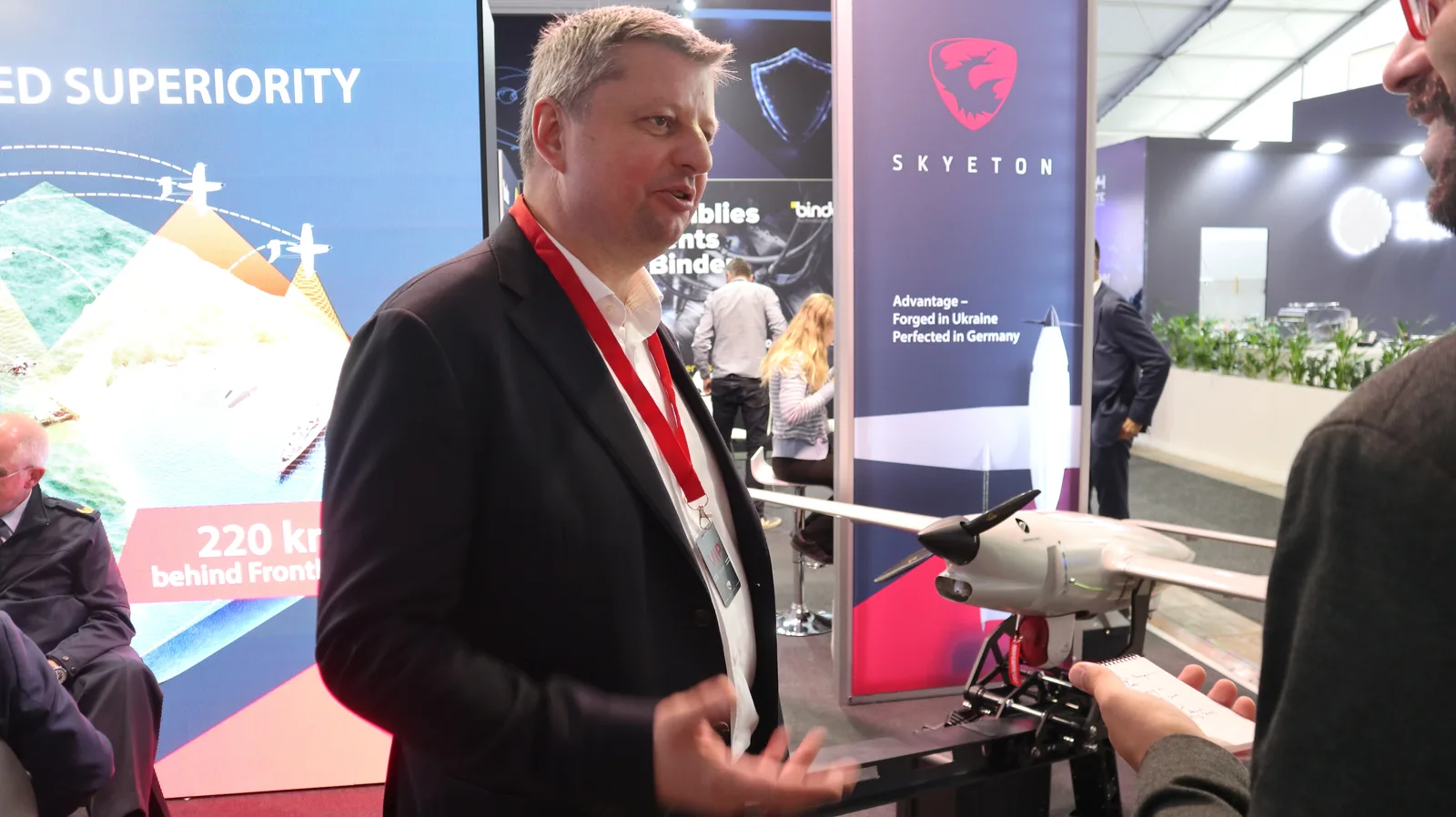 Ein Mann im Anzug führt auf einer Fachmesse ein Gespräch mit einer Person, die sich Notizen auf einem Block macht. Im Hintergrund sind Informationswände eines Herstellers von unbemannten Fluggeräten sowie das Modell einer kleinen Drohne mit Propellerantrieb auf einem Ausstellungsstand abgebildet.