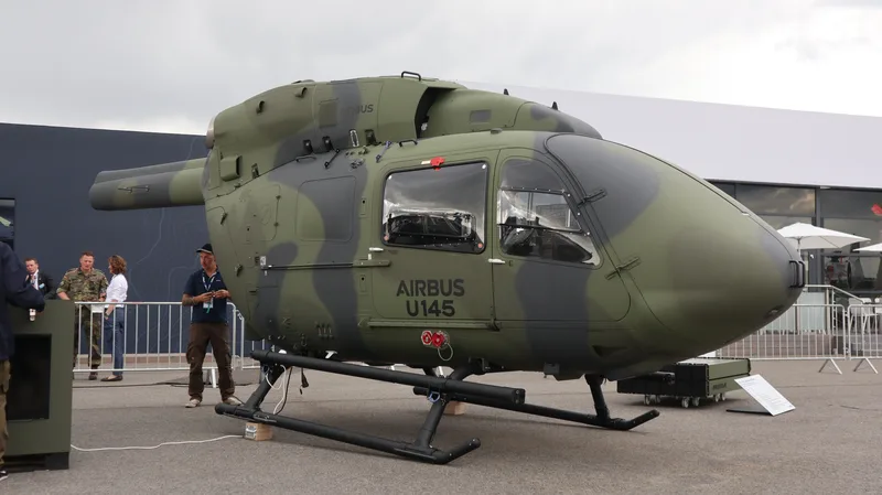 Auf einer Freifläche ist ein militärischer Hubschrauber mit Tarnanstrich sowie Hersteller- und Modellbezeichnung ausgestellt. Im Hintergrund stehen mehrere Personen, darunter ein Soldat in Flecktarn-Uniform, hinter einer Absperrung vor einem Ausstellungsgebäude.