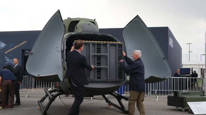 Zwei Männer präsentieren auf dem Außengelände einer Fachmesse eine modulare Startvorrichtung im geöffneten Heck eines militärischen Hubschraubers. Im Hintergrund sind Ausstellungsbauten eines Luftfahrtkonzerns sowie eine Informationstafel und eine Transportkiste eines Rüstungsunternehmens zu sehen.