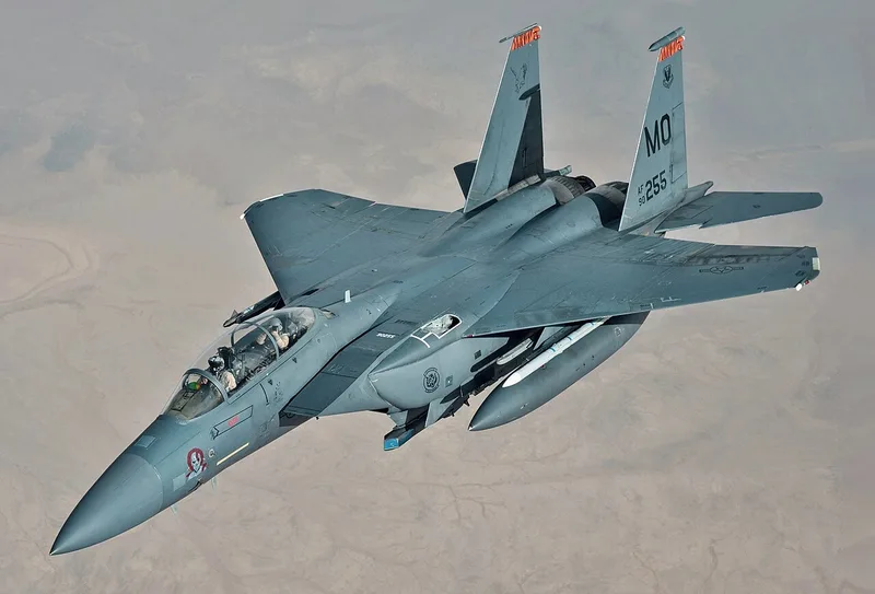 Ein zweisitziges Mehrzweckkampfflugzeug des Typs McDonnell Douglas F-15E Strike Eagle der US-Luftwaffe fliegt über einer Wüstenlandschaft. Die geöffnete Tanköffnung auf der linken Rumpfoberseite hinter dem Cockpit weist auf eine Luftbetankung hin.