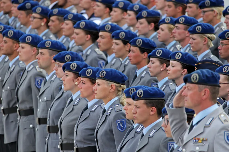 Soldatinnen und Soldaten des Sanitätsdienstes der Bundeswehr stehen in Formation bei einem feierlichen Gelöbnis. Sie tragen den grauen Dienstanzug mit dem blauen Barett und dem Abzeichen des Sanitätsdienstes, während ein Soldat im Vordergrund salutiert.