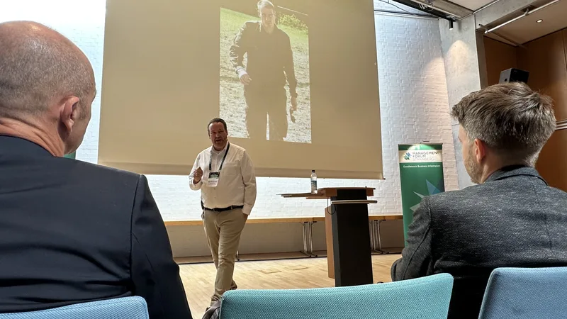 Ein Referent hält vor sitzenden Zuhörern einen Vortrag auf einer Bühne, neben der ein Werbebanner des Management Forum Starnberg steht. Im Hintergrund zeigt eine Leinwand das Foto einer Person im Freien.