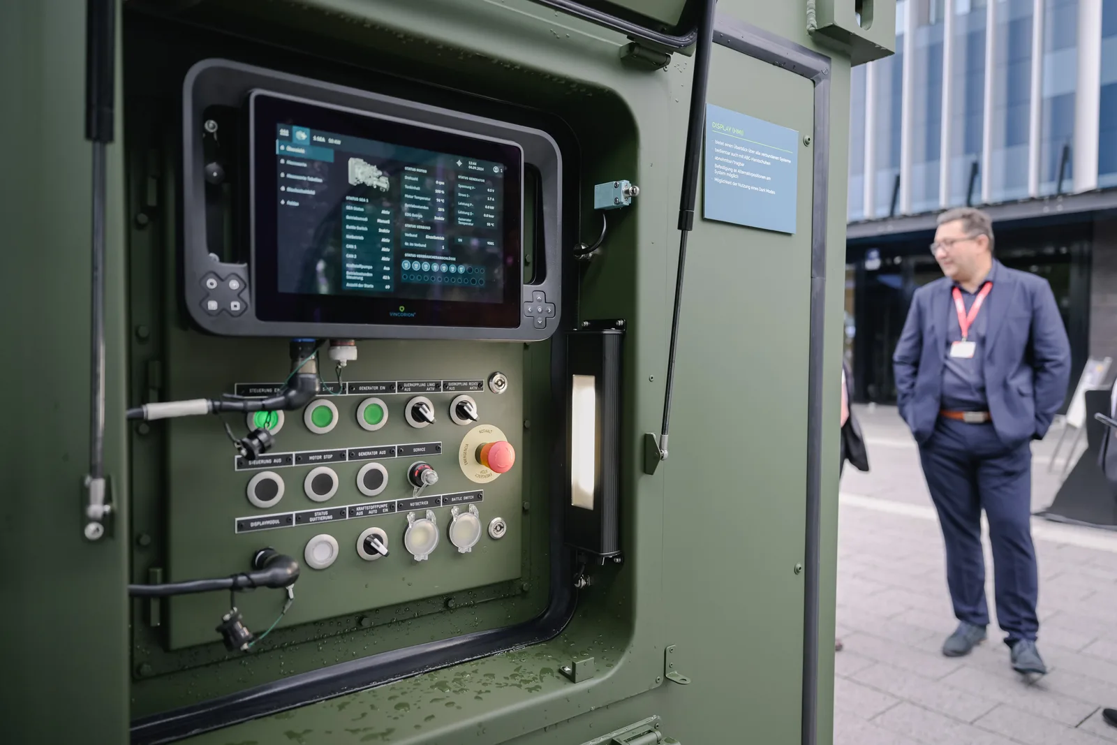 Die Steuerungseinheit eines militärischen Stromerzeugungsaggregats des Herstellers Vincorion zeigt einen robusten Bildschirm zur Systemüberwachung sowie diverse Schalter und einen roten Nothalt-Knopf. Im Hintergrund ist ein Besucher im Außenbereich einer Fachveranstaltung zu sehen.