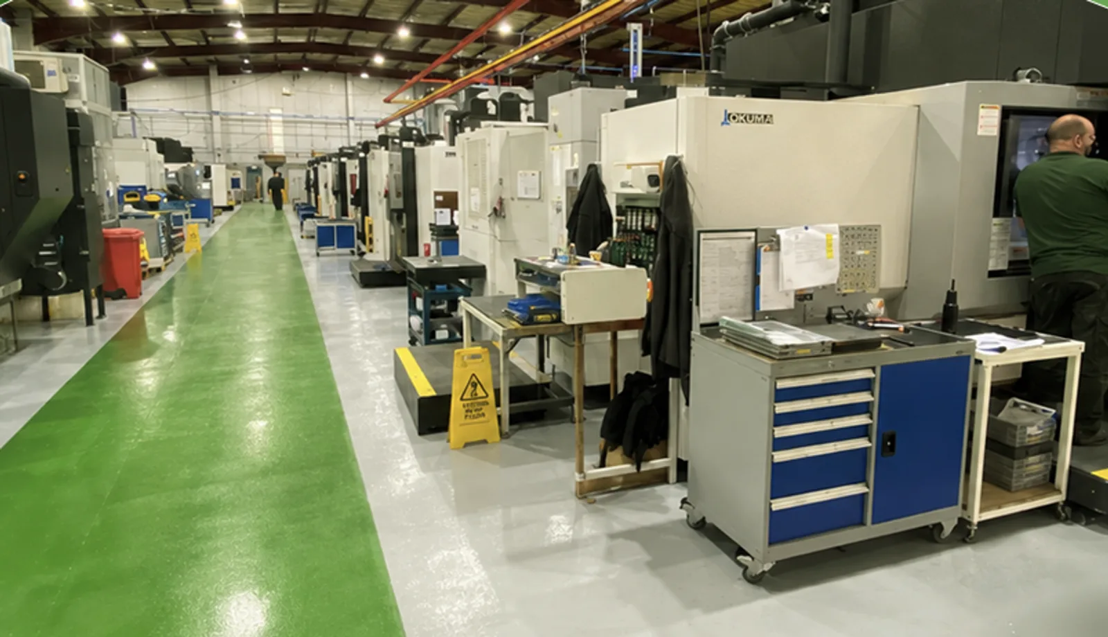 Blick in eine industrielle Fertigungshalle mit mehreren Werkzeugmaschinen, darunter Anlagen des Herstellers Okuma. Ein Mitarbeiter arbeitet an einer der Maschinen auf der rechten Seite, während ein grün markierter Gehweg durch die Halle führt.