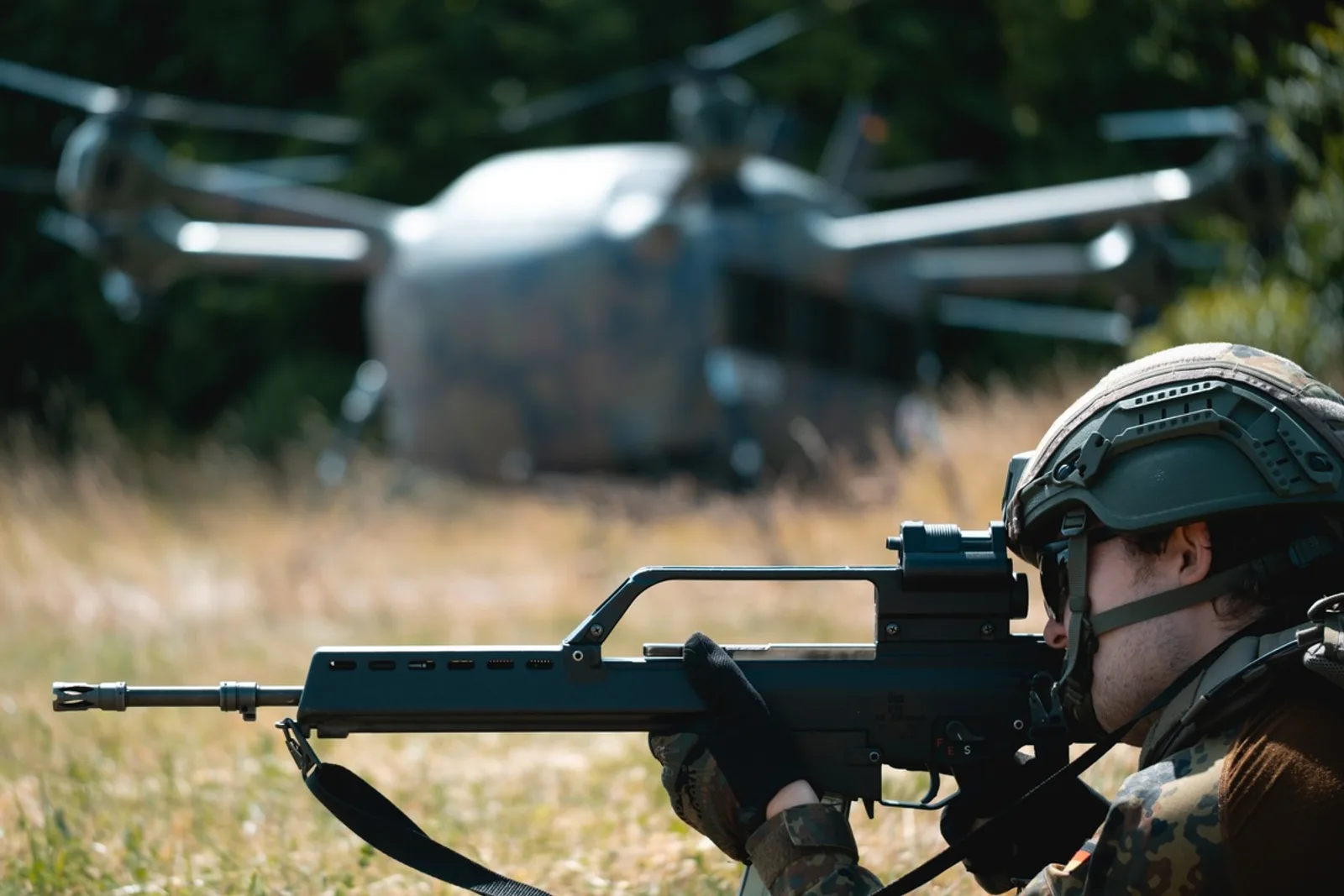 Ein Soldat sichert mit einem Sturmgewehr des Typs Heckler & Koch G36 ein Gelände. Im Hintergrund befindet sich die unbemannte Transportdrohne „Grille“ auf einer Freifläche.