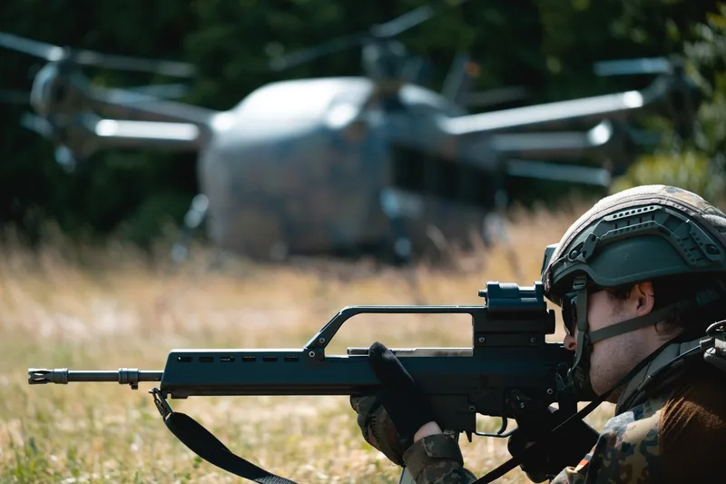 Ein Soldat sichert mit einem Sturmgewehr des Typs Heckler & Koch G36 ein Gelände. Im Hintergrund befindet sich die unbemannte Transportdrohne „Grille“ auf einer Freifläche.