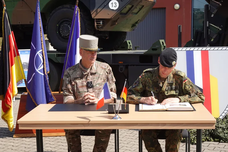 Generalmajor Christian Freuding und General Pierre Schill an einem Tisch mit Flaggen und einem französischen VAB-Transportpanzer