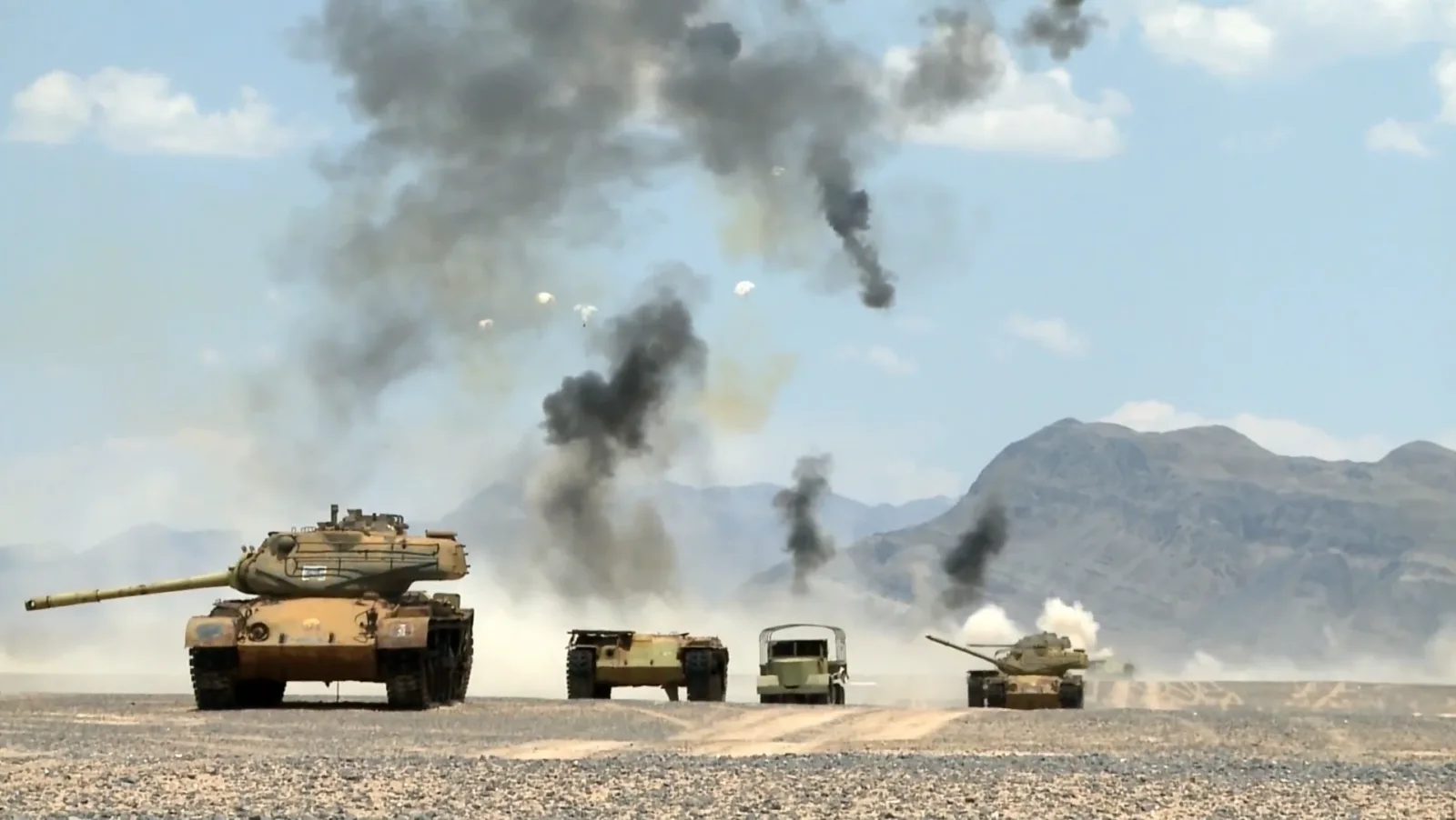 Ausgemusterte Militärfahrzeuge, darunter Kampfpanzer des Typs M47 Patton, dienen als Ziele bei einem Test von Streumunition der US-Luftwaffe. Über dem Wüstengelände sind herabsinkende Submunitionen an Fallschirmen sowie dichte Rauchwolken von Detonationen zu sehen.