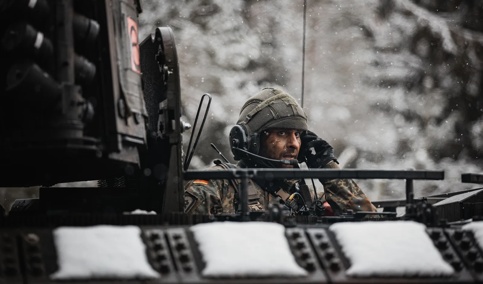 Ein Soldat der Bundeswehr blickt bei winterlichen Bedingungen aus der Luke eines Schützenpanzers Puma. Er trägt eine Sprechhaube zur Kommunikation, während im Vordergrund die charakteristische, schneebedeckte Dachpanzerung des Kettenfahrzeugs zu sehen ist.