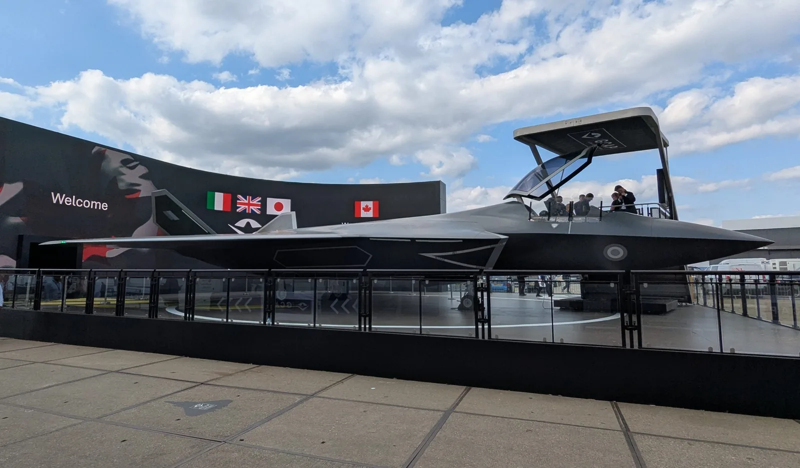 Das GCAP-Modell in Originalgröße vor dem Edgewing-Gebäude auf der Farnborough International Airshow 2026, mit der kanadischen Flagge neben denen Großbritanniens, Italiens und Japans auf der dahinterliegenden elektronischen Anzeigetafel, da Kanada Beobachterstatus im Programm erhält.