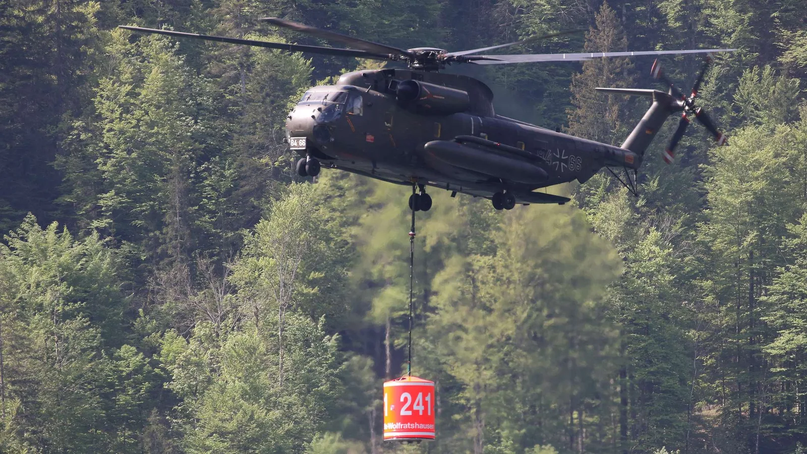 Ein Transporthubschrauber des Typs Sikorsky CH-53 der Bundeswehr fliegt mit einem angehängten Löschwasserbehälter über ein dichtes Waldgebiet. Die Besatzung unterstützt mit dieser Außenlast die Brandbekämpfung aus der Luft.