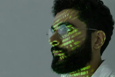 Ein Mann mit Brille und Bart im Profil, auf dessen Gesicht und Hals grüne Binärzeichen projiziert werden. Die Aufnahme visualisiert symbolisch die Themen der IT-Sicherheit und der digitalen Identifikation.