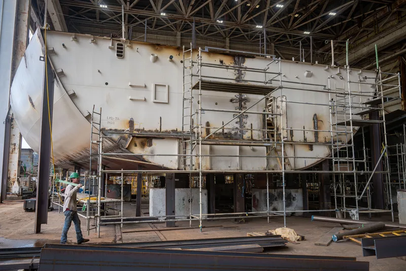 Werftarbeiter mit grünem Schutzhelm vor dem im Bau befindlichen Schiffsrumpf der LPD-32 in einer Schiffbauhalle