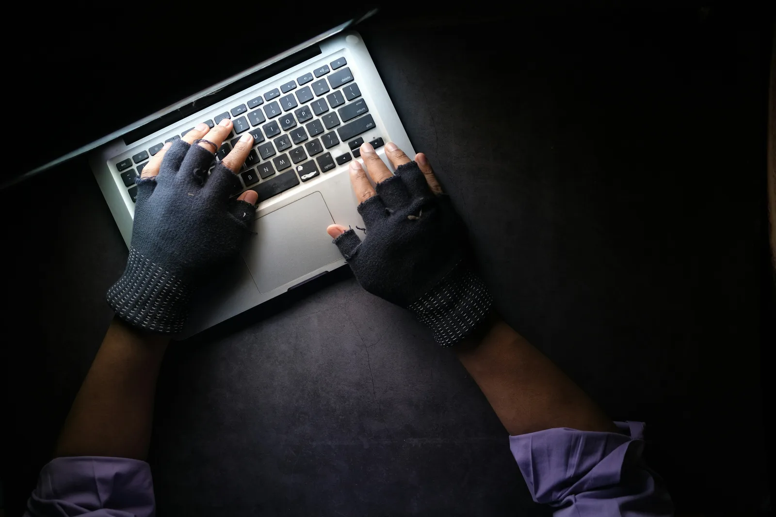 Eine Person mit fingerlosen, schwarzen Handschuhen bedient die Tastatur eines silbernen Laptops auf einer dunklen Arbeitsfläche. Die Aufnahme aus der Vogelperspektive zeigt die Hände und Unterarme des Nutzers im Schein des Bildschirms.