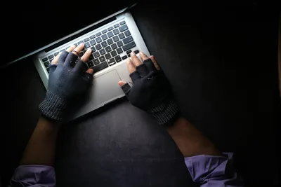 Eine Person mit fingerlosen, schwarzen Handschuhen bedient die Tastatur eines silbernen Laptops auf einer dunklen Arbeitsfläche. Die Aufnahme aus der Vogelperspektive zeigt die Hände und Unterarme des Nutzers im Schein des Bildschirms.