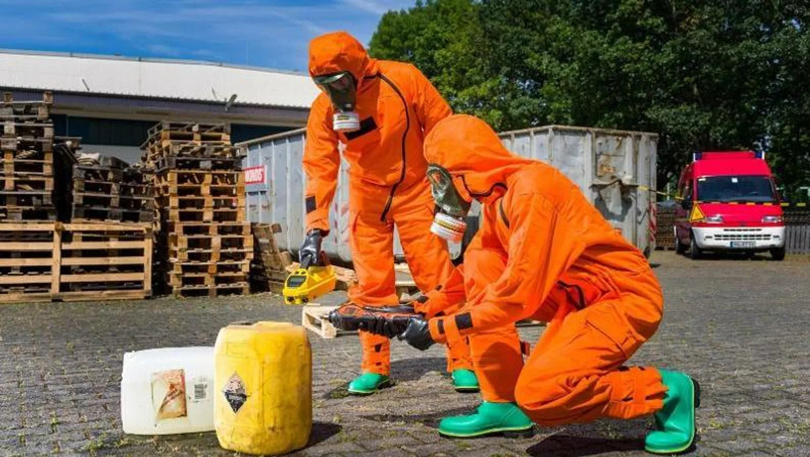 Zwei Einsatzkräfte in orangefarbenen Chemikalienschutzanzügen und Atemschutzmasken untersuchen mit mobilen Messgeräten zwei auf dem Boden stehende Kanister. Im Hintergrund eines Industriegeländes sind Holzpaletten, ein Container sowie ein rotes Einsatzfahrzeug zu sehen.