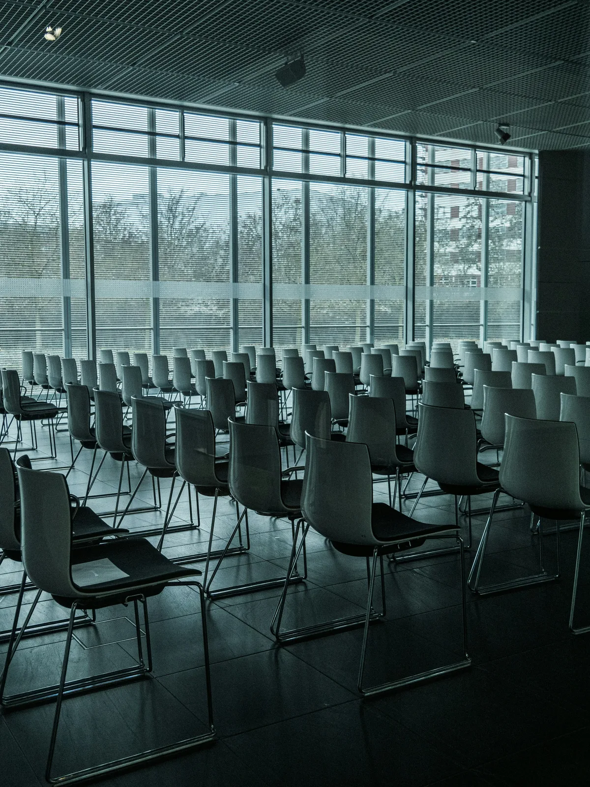 Reihen von leeren, modernen Stühlen stehen in einem Konferenz- oder Veranstaltungsraum vor einer großen Fensterfront mit Jalousien. Der Saal ist für eine Tagung oder Präsentation vorbereitet.